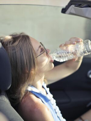 High angle side view of young woman in glasses and casual clothes drinking water from plastic transparent bottle while sitting in cabriolet with open roof in traffic jam in hot sunny day